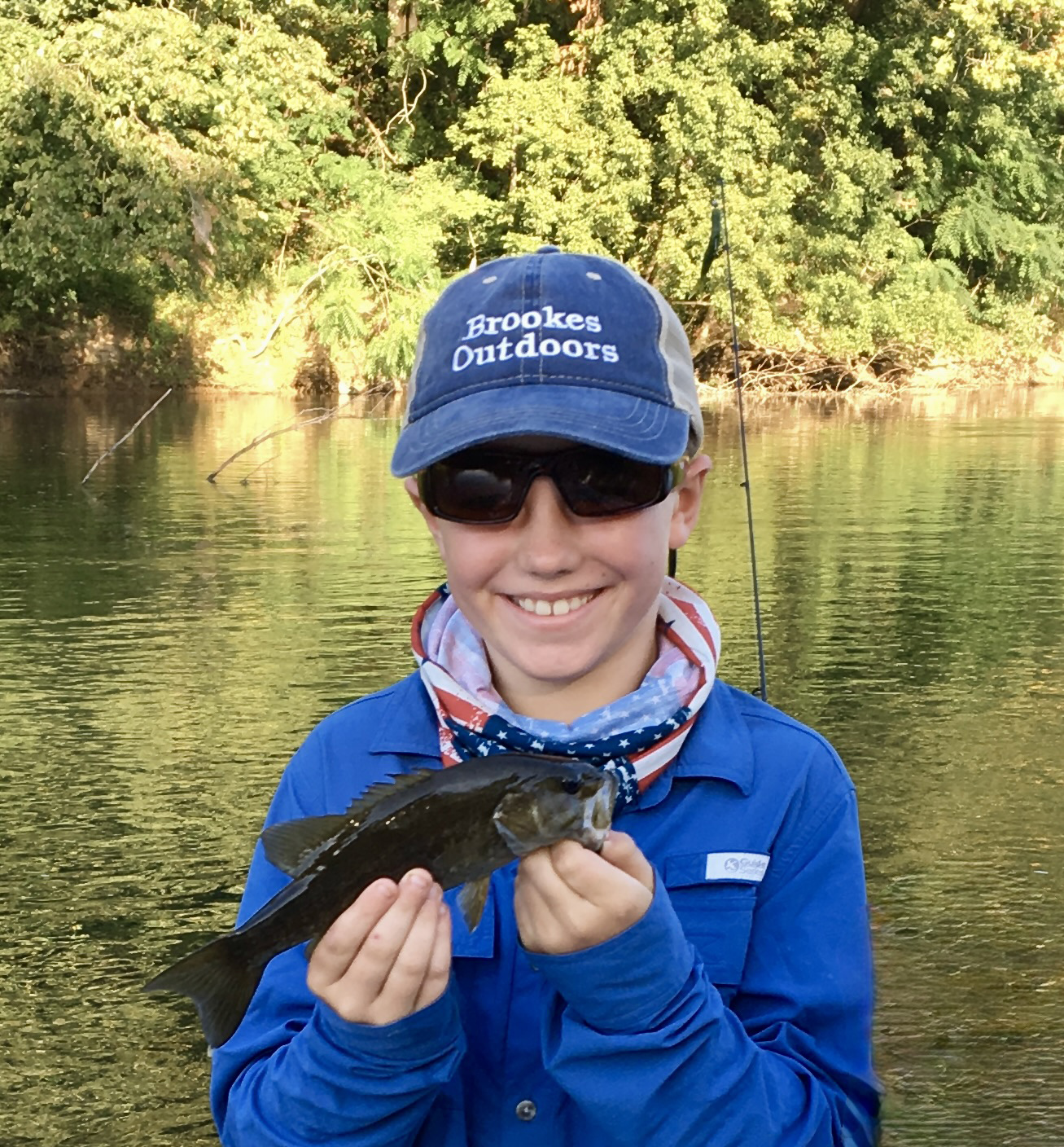 A photo of a boy holding up a smallmouth bass and smiling with a river behind him.